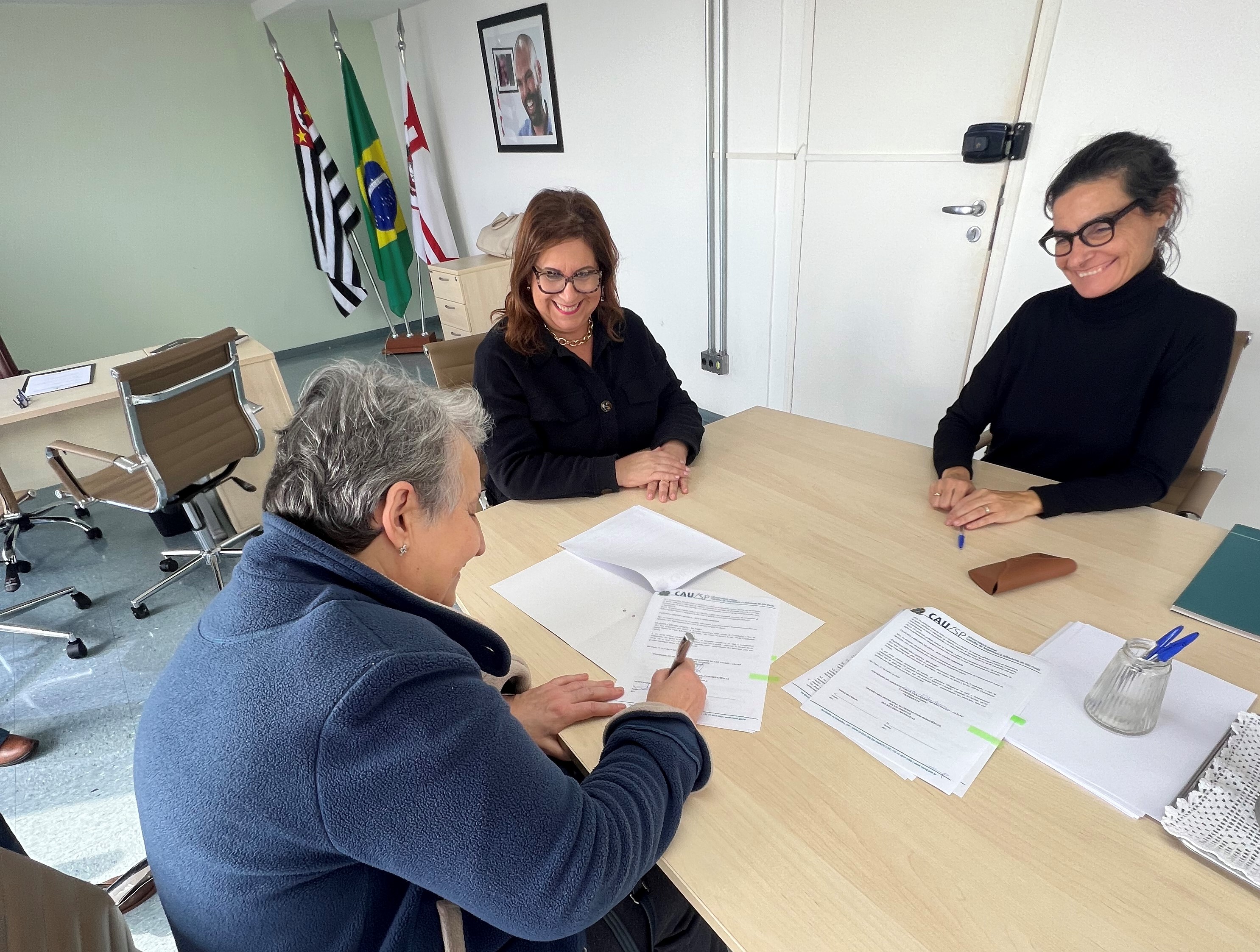 ao redor de uma mesa, no gabinete da SMPED, a secretária municipal da Pessoa com Deficiência Silvia Grecco, a presidente do CAU/SP Catherine Otondo e a Coordenadora da Comissão Temporária de Acessibilidade (CTA-CAU/SP), Maria Alice Gaiotto. Maria Alice Gaiotto assina o termo sobre a mesa.