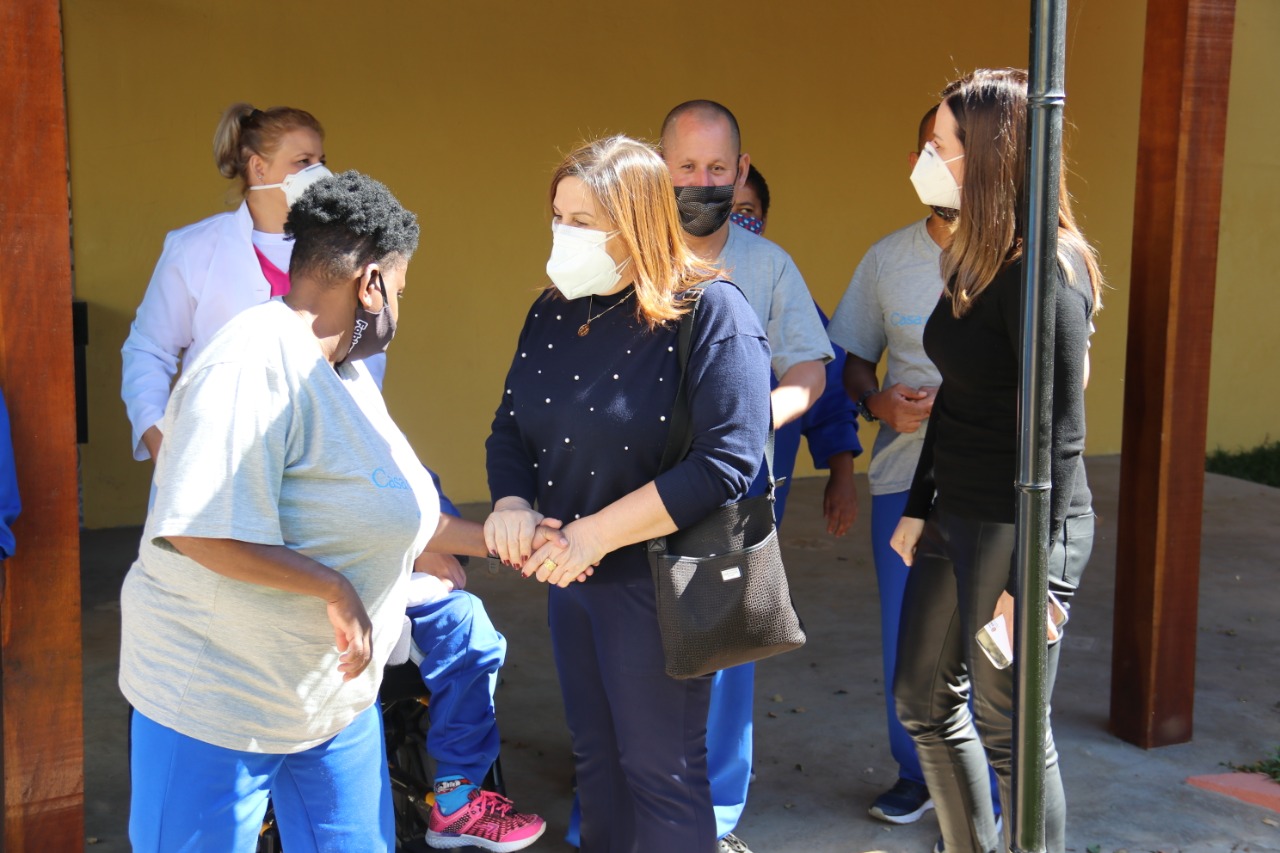 Do lado externo da Casa de Davi, secretária da SMPED, Silvia Grecco cumprimenta os pacientes do local com a Claudia Carletto, Secretária Municipal de Direitos Humanos e Cidadania. Todos usam máscaras de proteção.