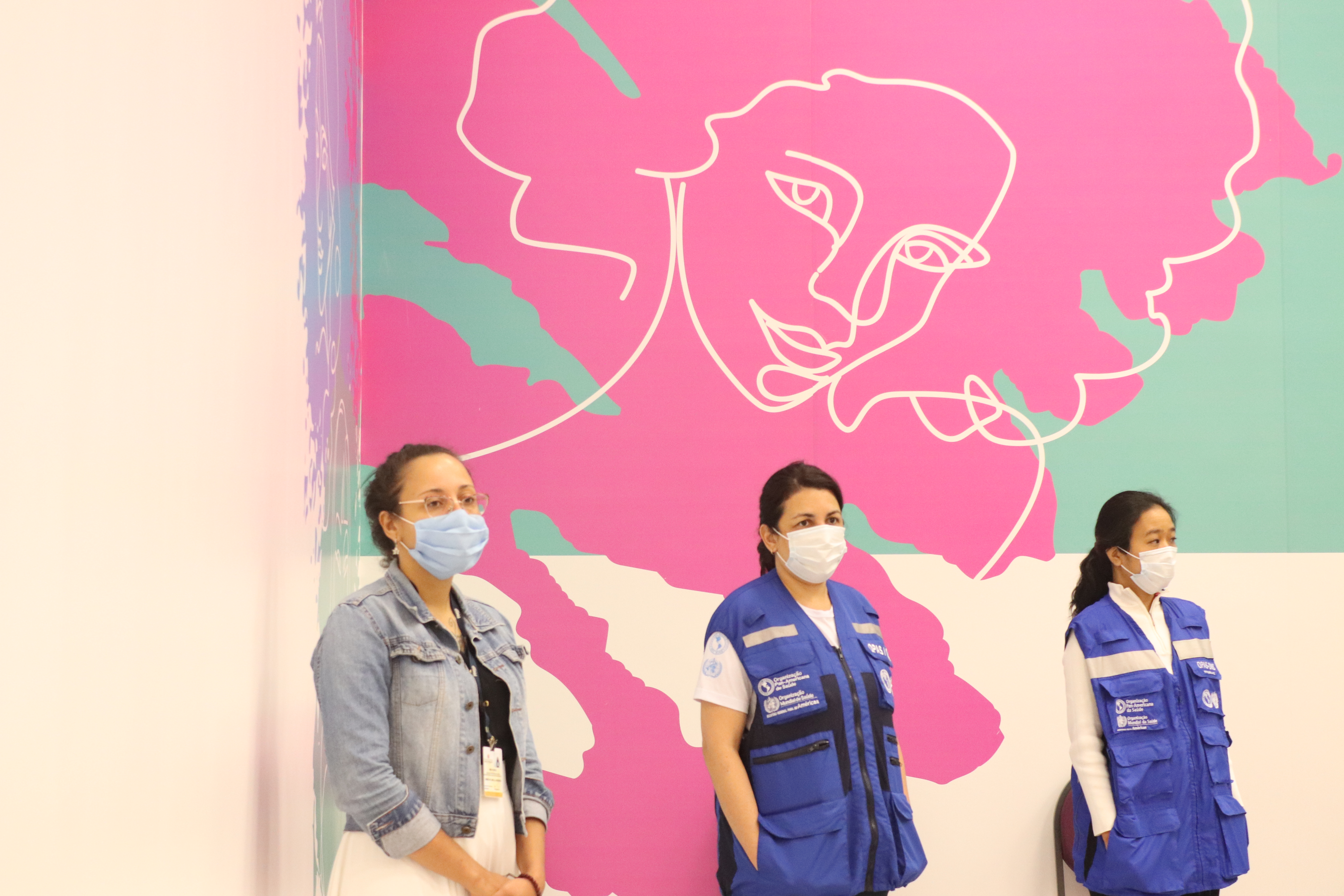 Foto da capacitação com três mulheres em pé assistindo à palestra; no fundo há um painel cor de rosa com um contorno de um rosto de mulher. 