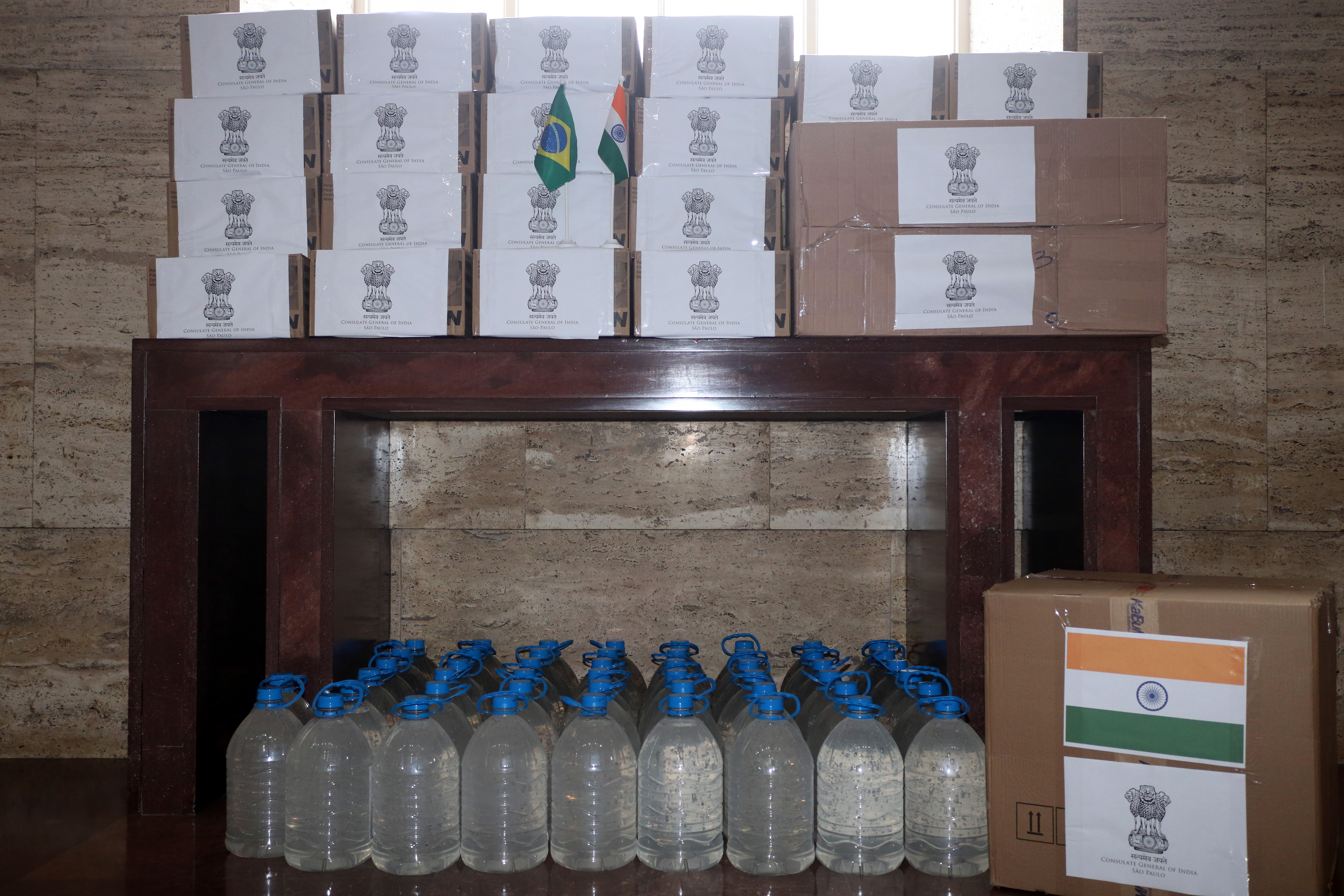 Foto dos produtos doados. Em cima de uma mesa estão caixas com a Bandeira da Índia do Brasil; embaixo na mesa, estão os galões de álcool em gel e, ao lado dos galões, mais caixas de doações. 