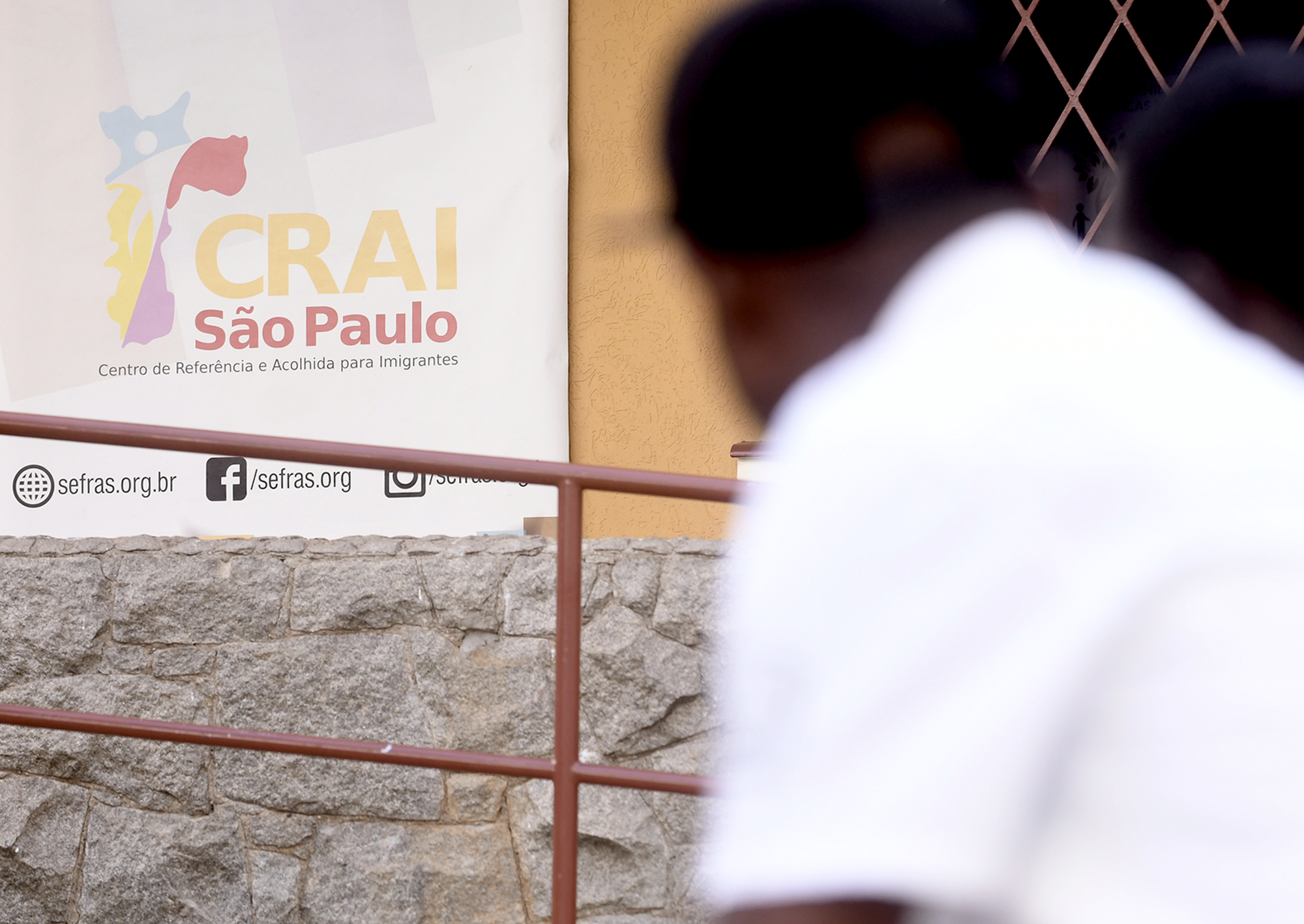 Foto da entrada de um Centro de Referência e Acolhida para Imigrantes (CRAI) com um homem negro em primeiro plano, desfocado, à direita na imagem. 