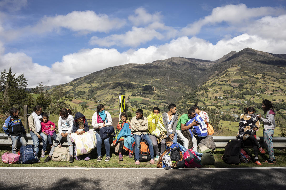 A foto mostra 16 venezuelanos, entre adultos e crianças, sentados a margem de uma estrada na Colômbia, com montanhas ao fundo. 