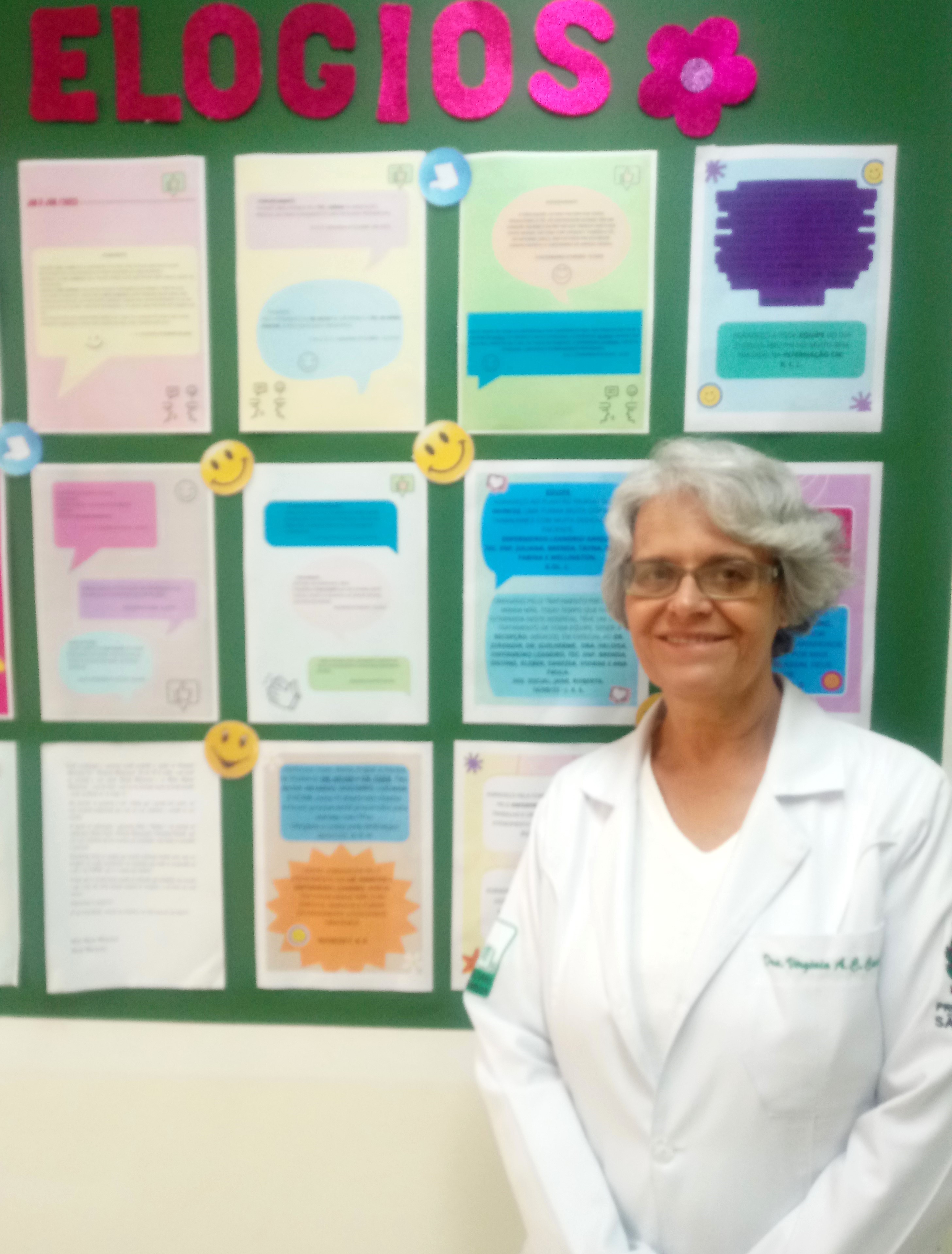A foto mostra a médica Virgínia Affonso em frente a um quadro com vários papéis afixados, trazendo impressões em várias cores; na parte de cima, em letras recortadas em cor-de-rosa, lê-se 