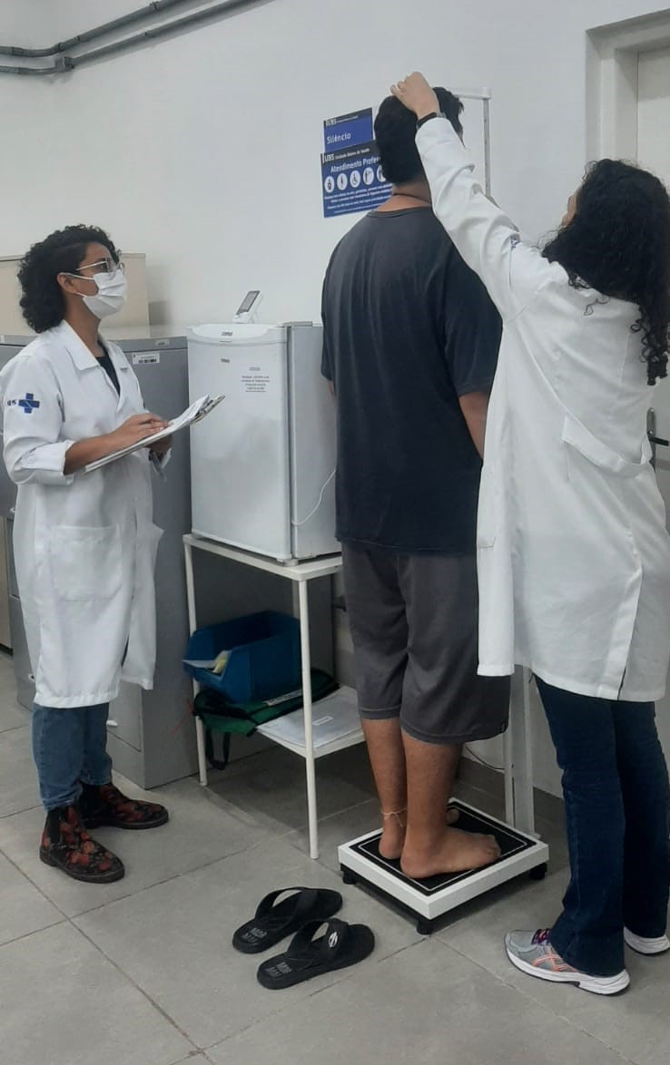 Na foto, duas profissionais de enfermagem, ambas usando jalecos e máscaras, realizam pesagem e medição de um homem jovem, ele está de costas, posicionado sobre uma balança