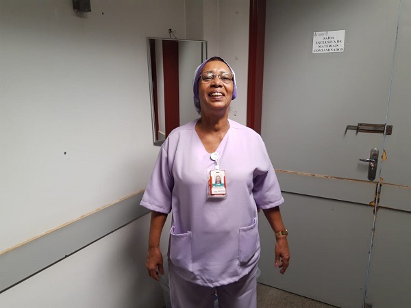  #PraCegoVer: Arlete está em pé e sorri, numa sala do hospital no qual trabalha. Ela usa roupa clara e óculos.
