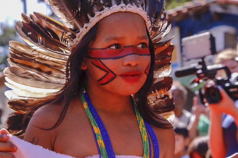 A foto mostra uma menina indígena usando cocar de penas, pintura no rosto e um colar de miçangas coloridas 