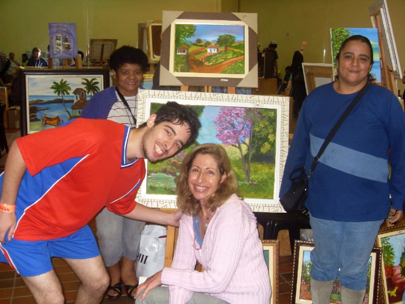 Benedita aparece mais ao fundo e, na frente dela, estão alguns quadros pintados pelos pacientes do Caps. Na foto estão ainda mais três pessoas, todos sorrindo