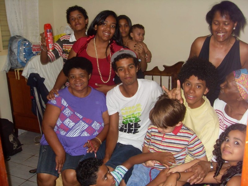 Benedita está na foto com parte dos filhos, netos e sobrinhos. Eles estão sentados sobre uma cama, sorrindo