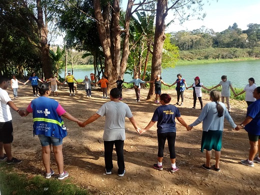 Um grupo de aproximadamente 30 pessoas está em pé, de mãos dadas, formando um círculo, ao ar livre. ao fundo é possível observar um lago.