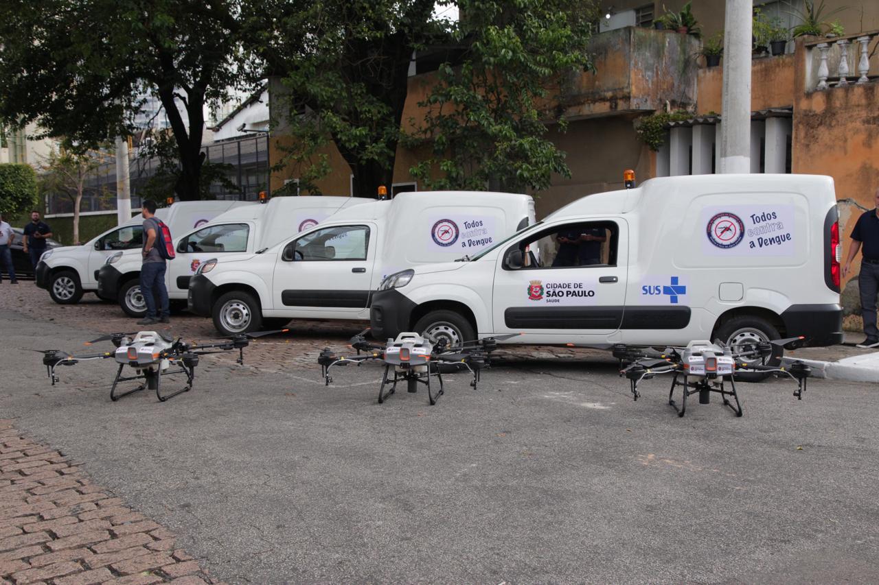 Na imagem, há quatro veículos brancos da Saúde estacionados em diagonal,e à frente tem três drones apoiados no chão