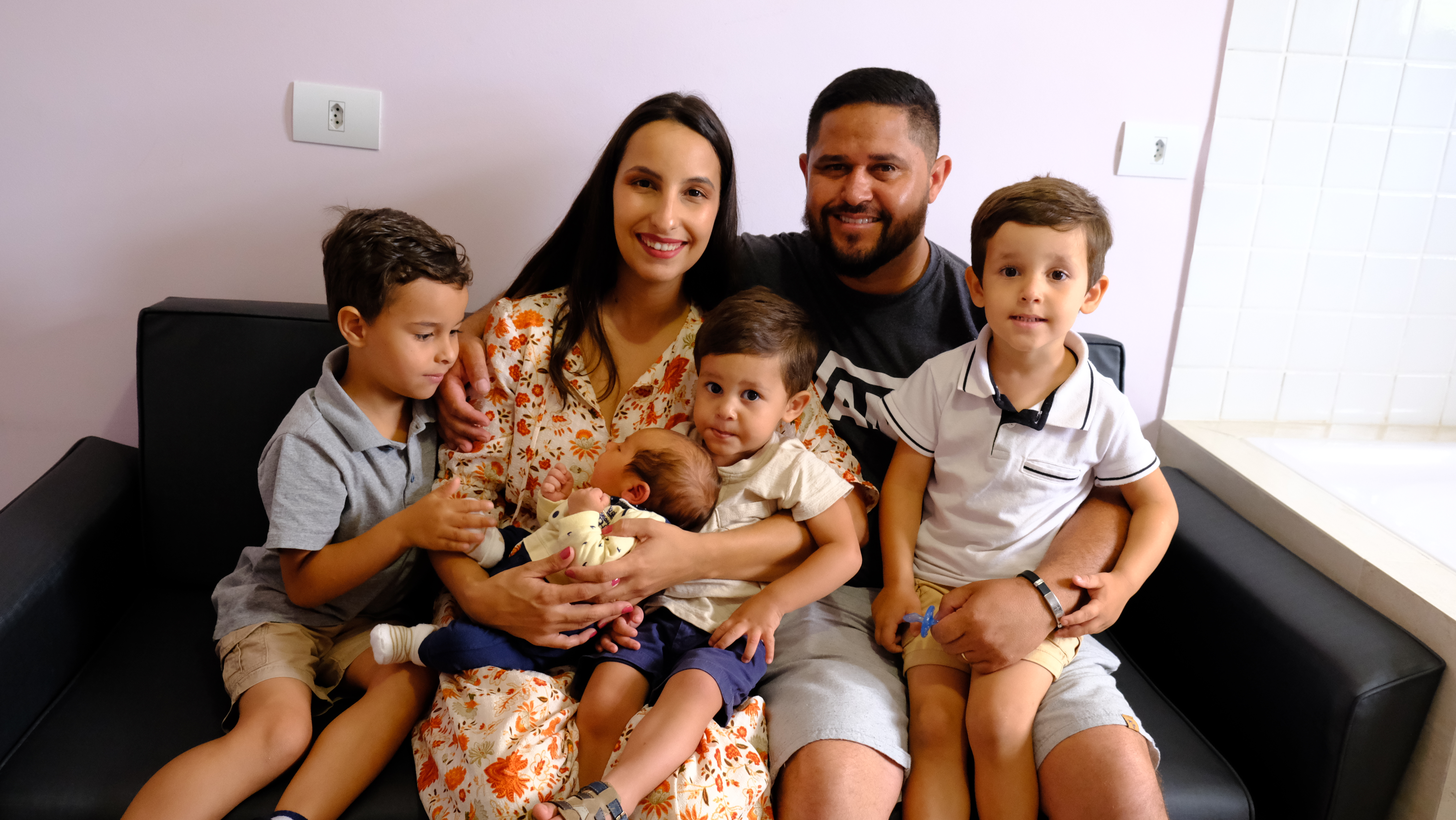 A foto mostra Bárbara, uma mulher branca, jovem, de cabelos pretos compridos. Ela está sentada em um sofá, segura um bebê no colo e está cercada de outras três crianças, todos meninos com menos de seis anos, e do marido, um homem moreno, de cabelos curtos e barba