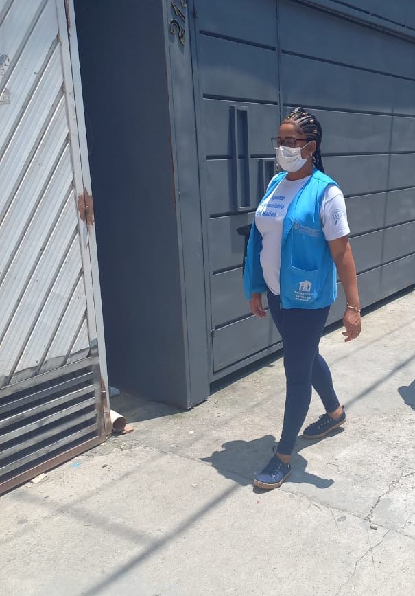 A foto mostra uma mulher negra de aparência jovial e cabelos trançados; ela usa máscara e um colete azul, e está caminhando na rua, em frente a uma residência 