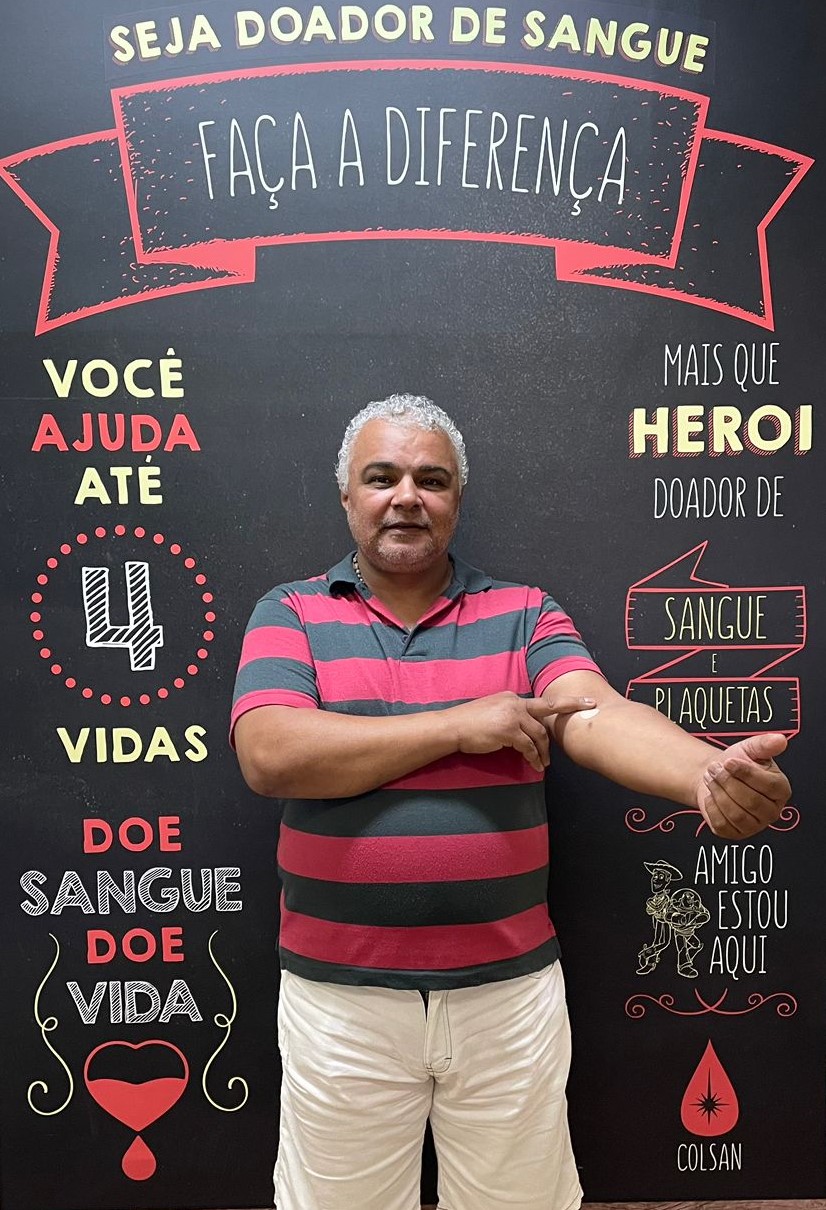 A foto mostra Antonio Roberto em frente a uma placa de incentivo à doação de sangue; ele é um homem de cabelos grisalhos  e está usando uma camiseta listrada de preto e vermelho; tem o braço esquerdo esticado, e com a mão direita aperta um pequeno curativo neste braço; na placa lê-se frases de icentivo à doação, como: 'Seja doador de sangue, faça a diferença', 'Doe sangue, doe vida' e outras  