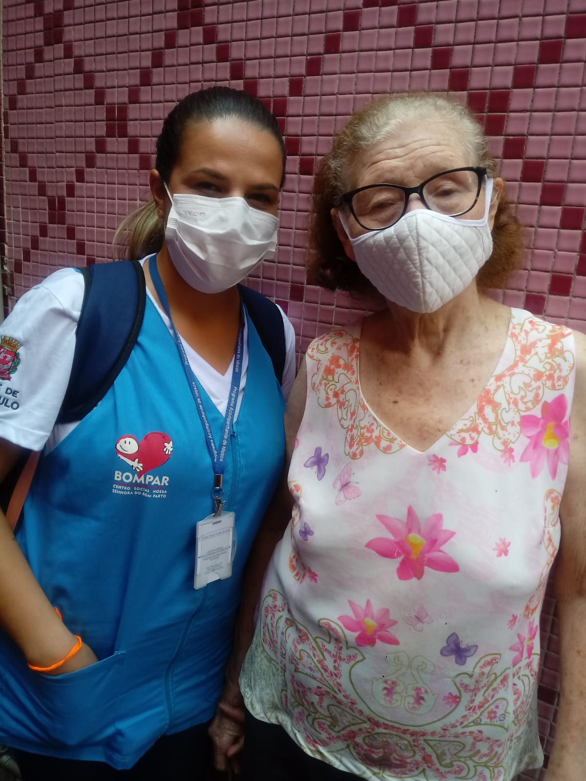 Gizelly posa com dona Lucília, uma mulher idosa que veste uma blusa com estampa de flores, em frente a uma parede revestida com pastilhas cor de vinho; ambas usam máscara 
