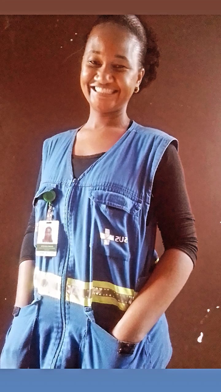 A foto mostra Duane, uma mulher negra e jovem, posando para a foto; ela usa um colete azul com a logomarca com Sistema Único de Saúde e também um crachá