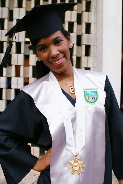Foto de Eliezka no dia de sua formatura. Ela está sorrindo para a foto vestida com uma beca preta e branca e capelo, chapéu típico de formandos, brincos e colar dourados. No pescoço ela usa uma fita branca com uma estrela grande pendurada