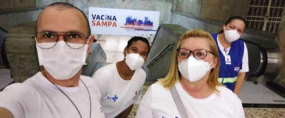 Eliezka está no centro da foto, atrás de duas pessoas e ao lado de uma terceira. Eles estão em frente a uma escada dentro de uma estação do metrô. Todos usam máscara branca e camiseta branca com o emblema do SUS