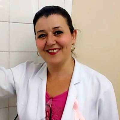 Foto de Elisabete Amador. De jaleco branco e blusa rosa, Elisabete sorri. Ela está em pé, próximo a uma parede bege de azulejos e tem os cabelos avermelhados presos em coque