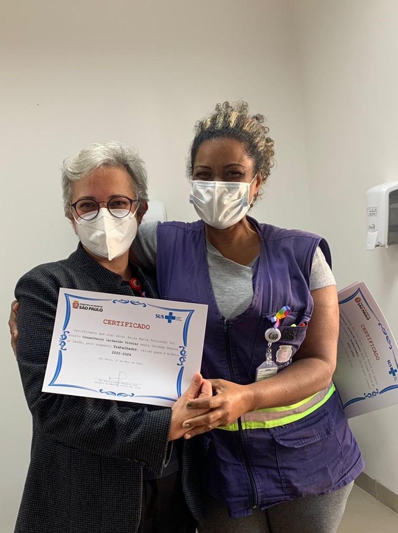 A foto mostra Erika, uma mulher negra, jovem, usando um colete azul e máscara; ela está abraçada a uma mulher branca, de cabelos brancos, de meia-idade, e ambas seguram um certificado, que está sendo entregue a Erika