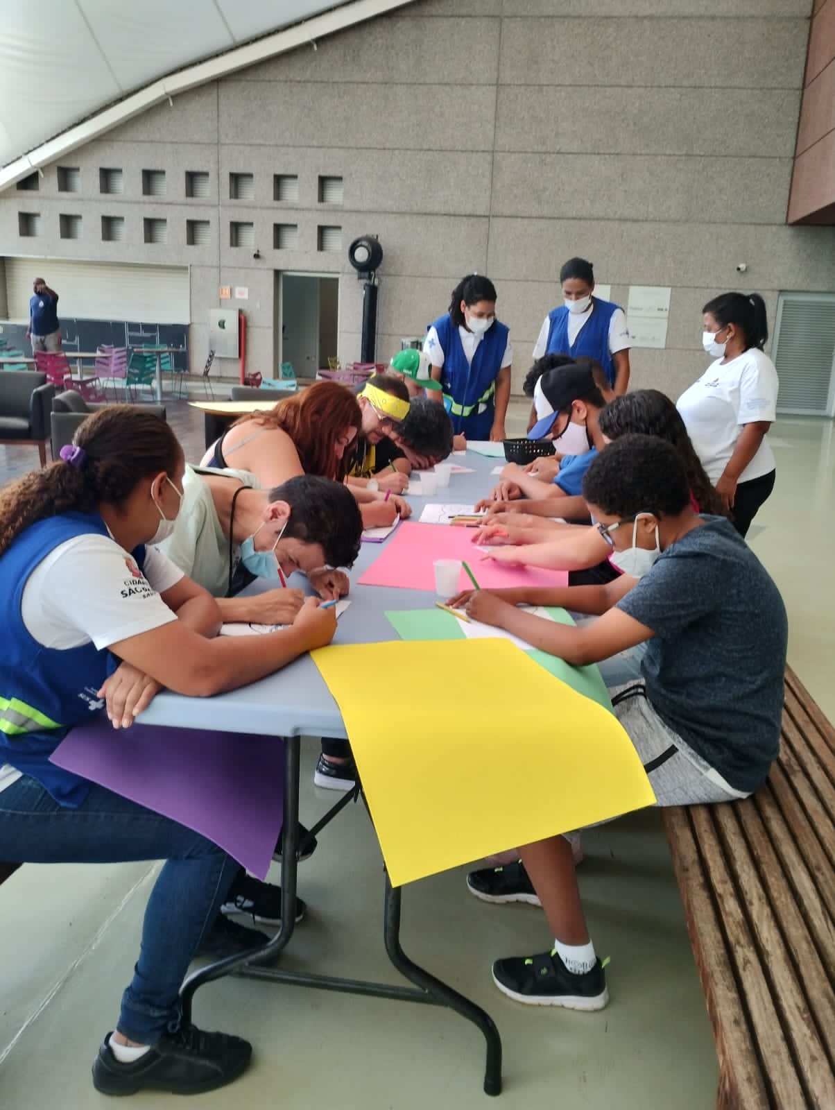 A foto mostra jovens sentados à volta de uma mesa grande; eles manipulam lápis de cor e folhas coloridas de cartolina