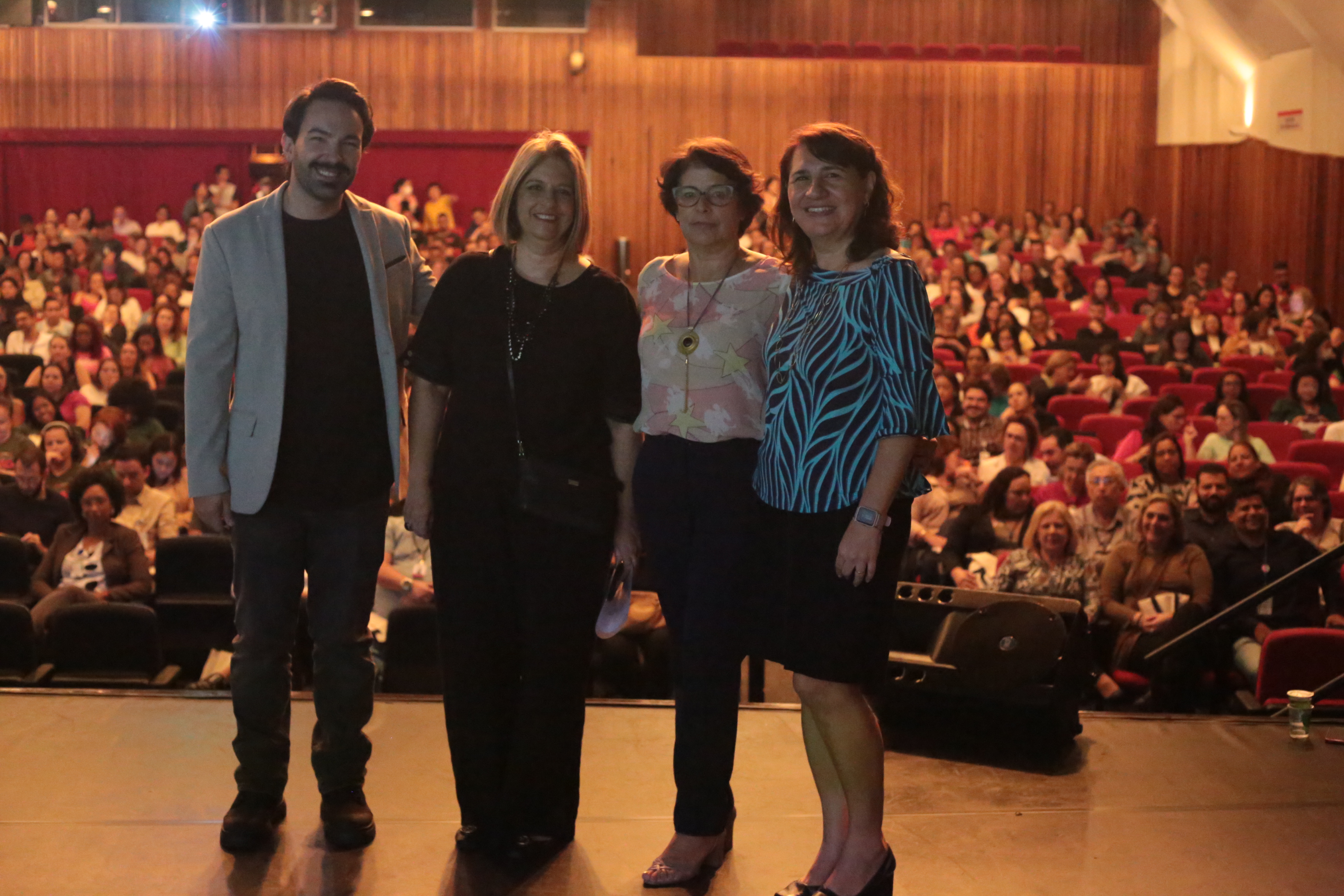 A foto mostra quatro pessoas, três mulheres e um homem, posando sobre um palco; às suas costas está uma plateia com centenas de pessoas sentadas