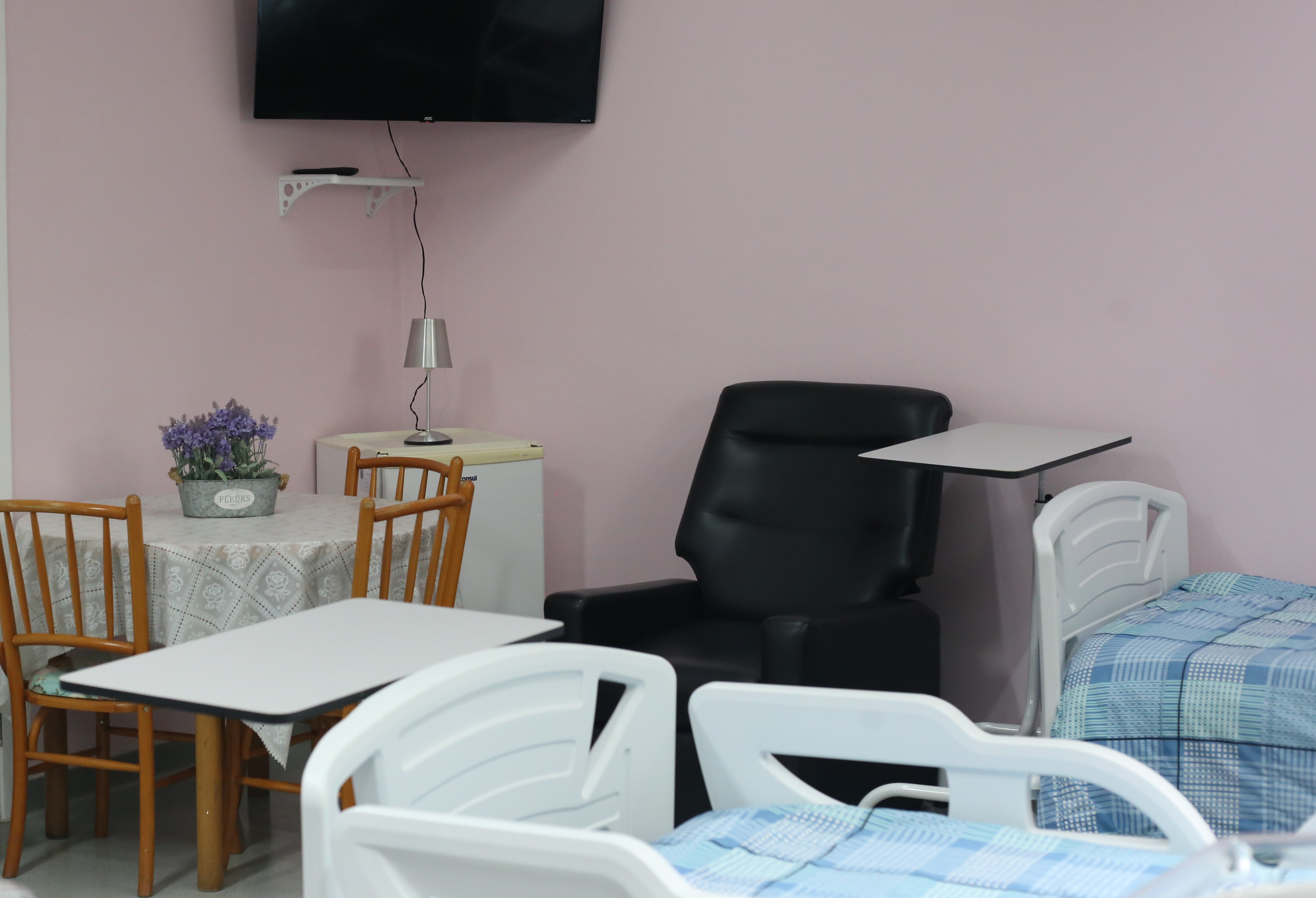A foto mostra uma suíte hospitalar; nela, vê-se duas camas com edredons em tons de azul, uma poltrona preta, além de uma mesa, frigobar e televisão