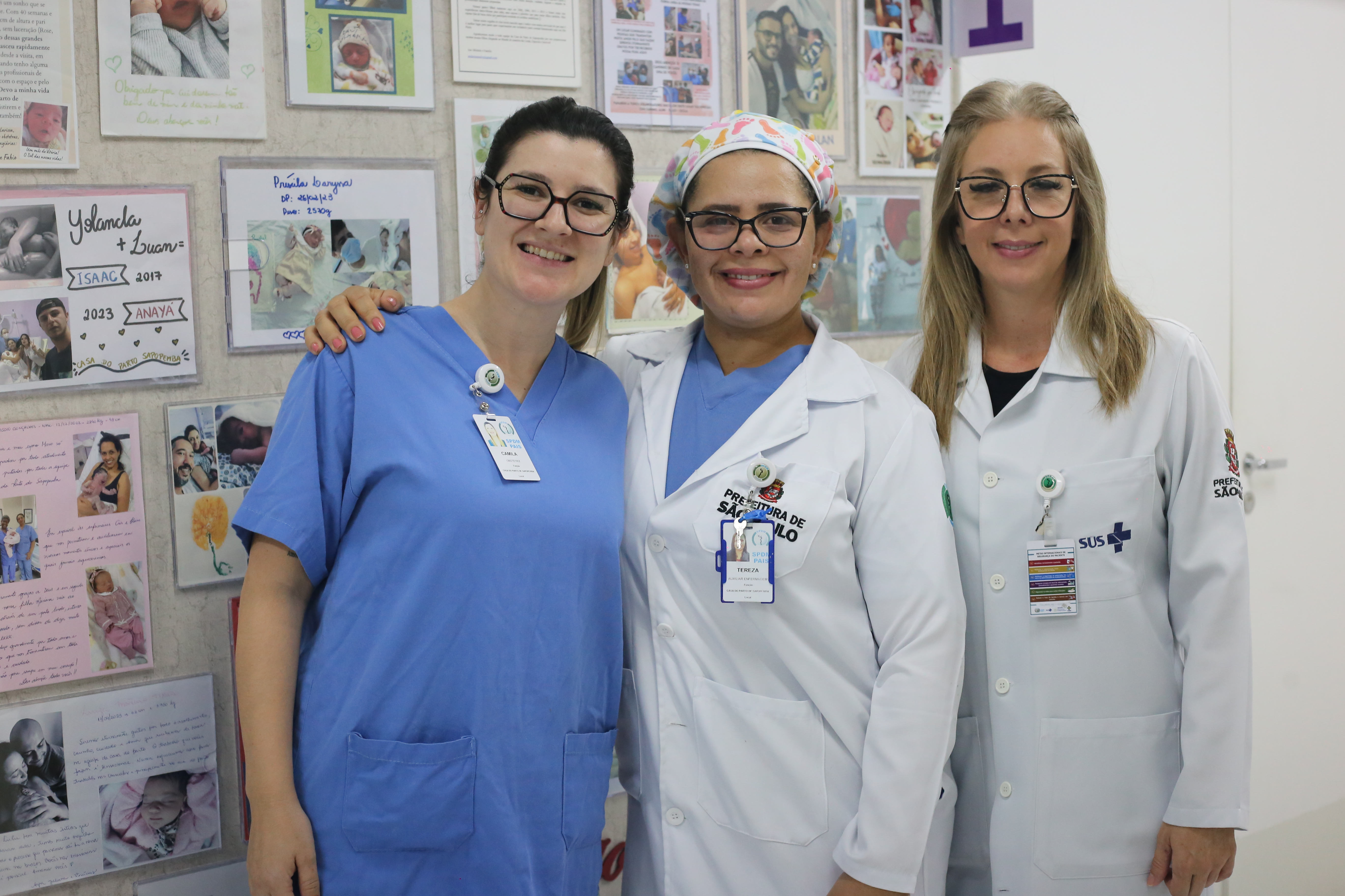 A foto mostra três mulheres posando em frente a um painel com fotos de bebês recém-nascidos; elas sorriem, duas delas usam jalecos brancos e uma veste um uniforme azul