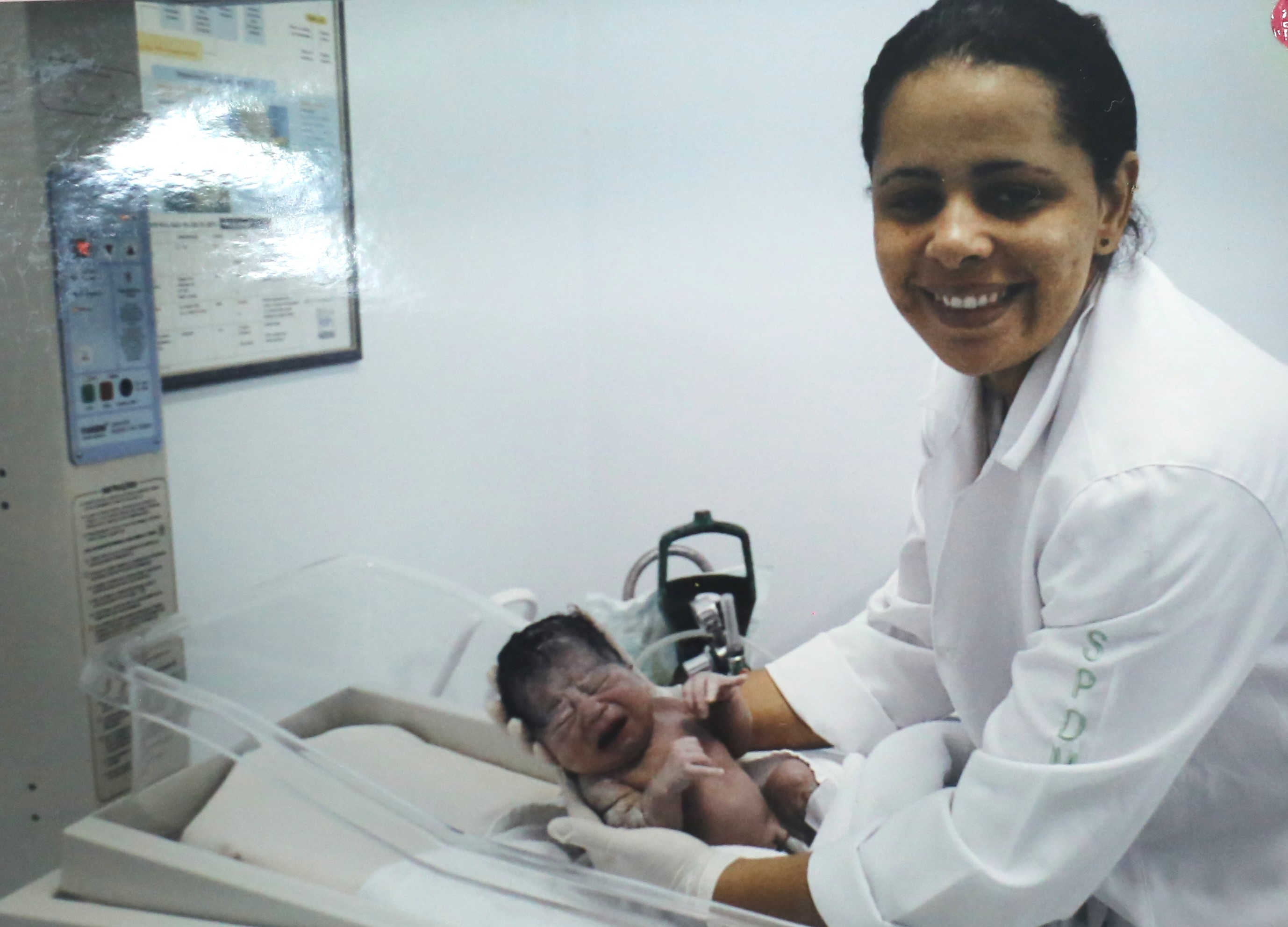 A foto mostra uma mulher jovem de cabelos pretos e presos; usando um jaleco branco; ela está inclinada sobre um berço hospitalar e segura um bebê recém-nascido, que chora