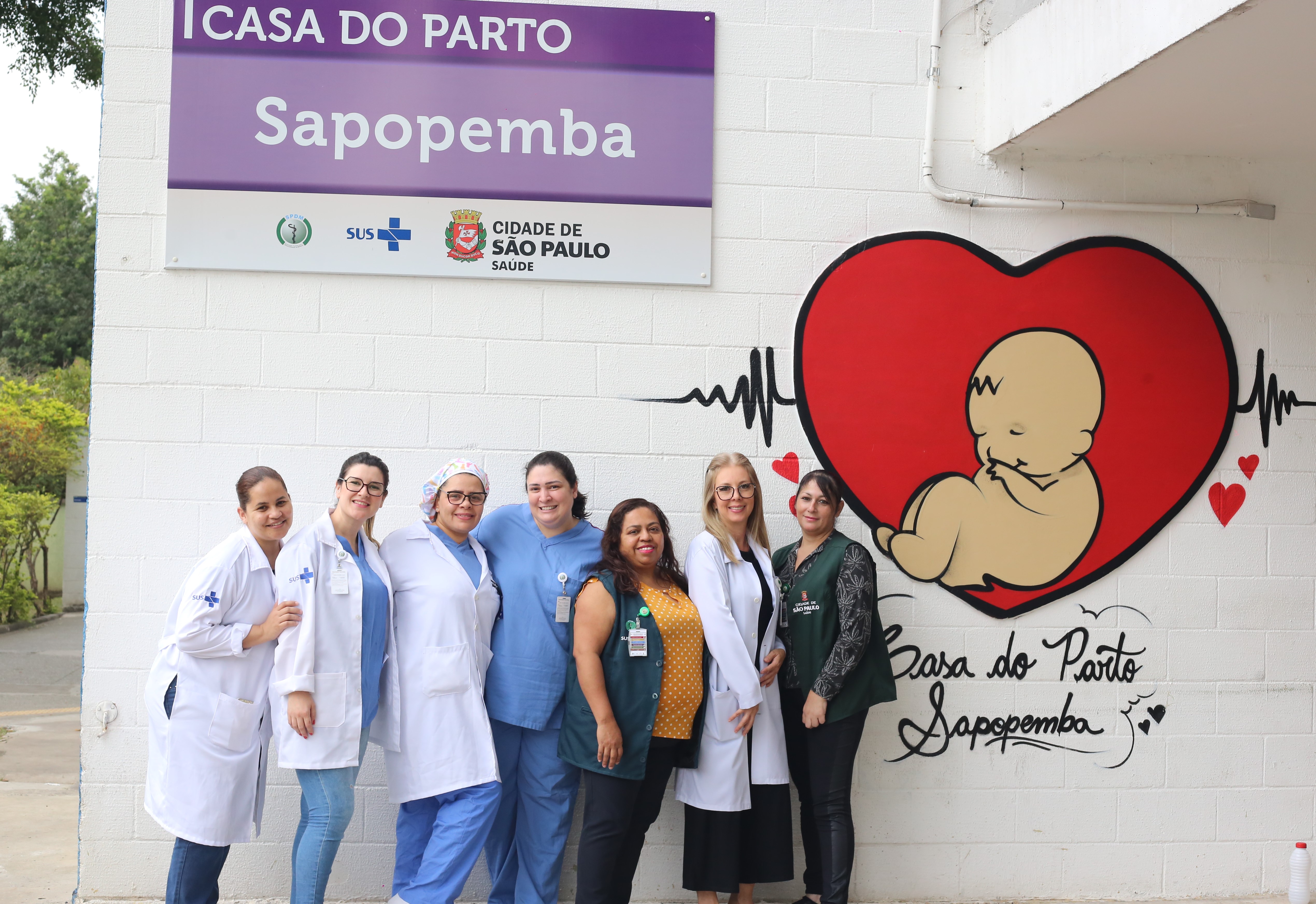 A foto mostra sete mulheres usando uniformes, posando em frente uma fachada, na qual estão uma placa identificando “Casa do Parto Sapopemba”, de um lado, e uma pintura mostrando um bebê recém-nascido dentro de um coração