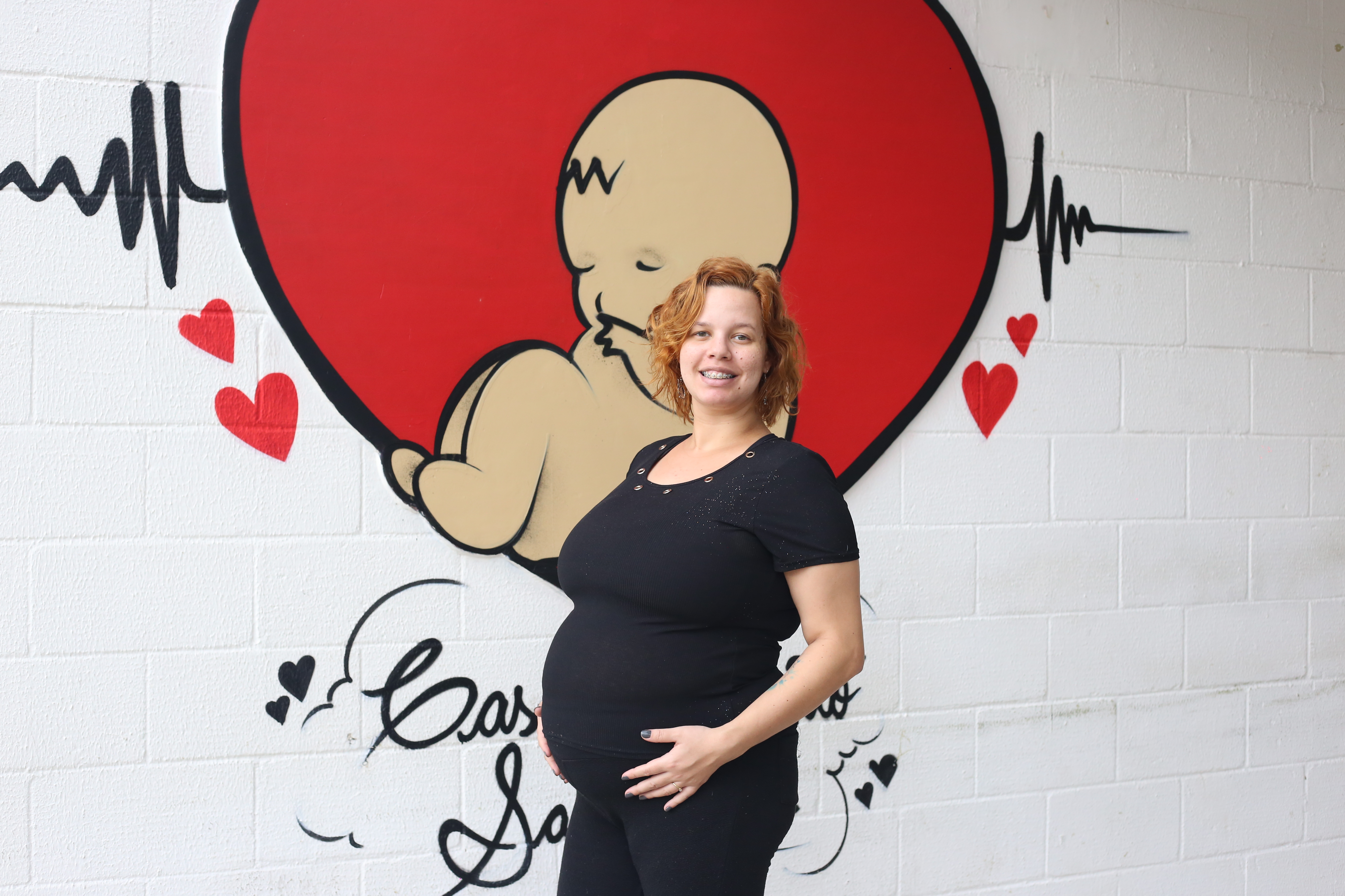 A foto mostra uma mulher jovem de cabelos ruivos; ela veste roupas pretas, está grávida e posa em frente a uma parede decorada com a pintura de um bebê dentro de um coração, que imita um útero