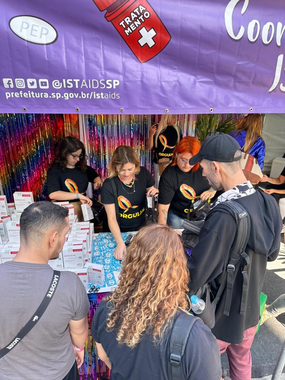 A foto mostra três pessoas da equipe de IST/Aids dentro de uma tenda; elas vestem camisetas pretas nas quais se lê a palavra "orgulho" e distribuem insumos a pessoas posicionadas do lado de fora da tenda
