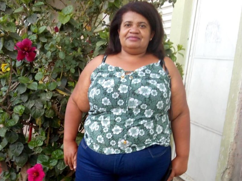 Foto de Janecleide antes da cirurgia. Ela posa na frente de uma planta e está usando uma blusa florida que deixa seus ombros de fora e esboça um leve sorriso