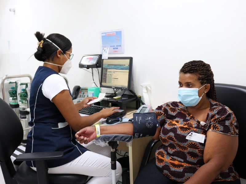 Foto de Janecleide durante uma consulta. Ela está sentada numa cadeira enquanto mede a pressão com um aparelho em seu braço. Ao seu lado, uma profissional da saúde está sentada de frente para um computador e digitando