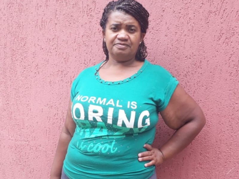 Foto de Janecleide depois da cirurgia. Ela está na frente de uma parede rosa usando uma blusa verde, olhando para a frente e com a mão na cintura. A boca esboça um leve sorriso