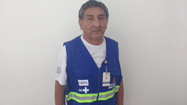 #PraCegoVer: Jorge está em pé e sorri. Usa o colete azul de Agente Comunitário de Saúde. Atrás dele há uma parede branca.