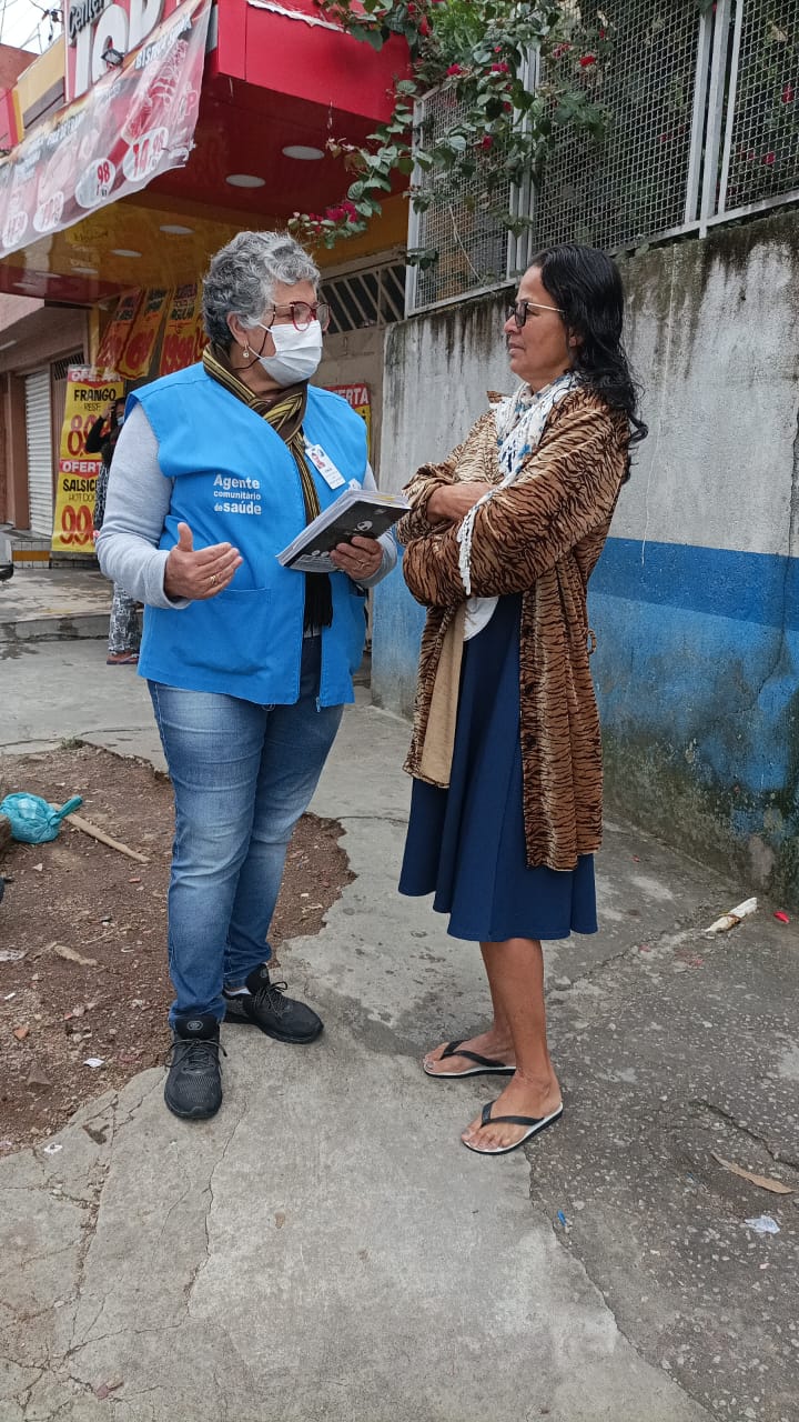 A foto mostra uma mulher branca, de meia-idade e cabelos brancos usando máscara e um colete azul; ela conversa com outra mulher, e ambas estão em um ambiente externo, uma calçada 