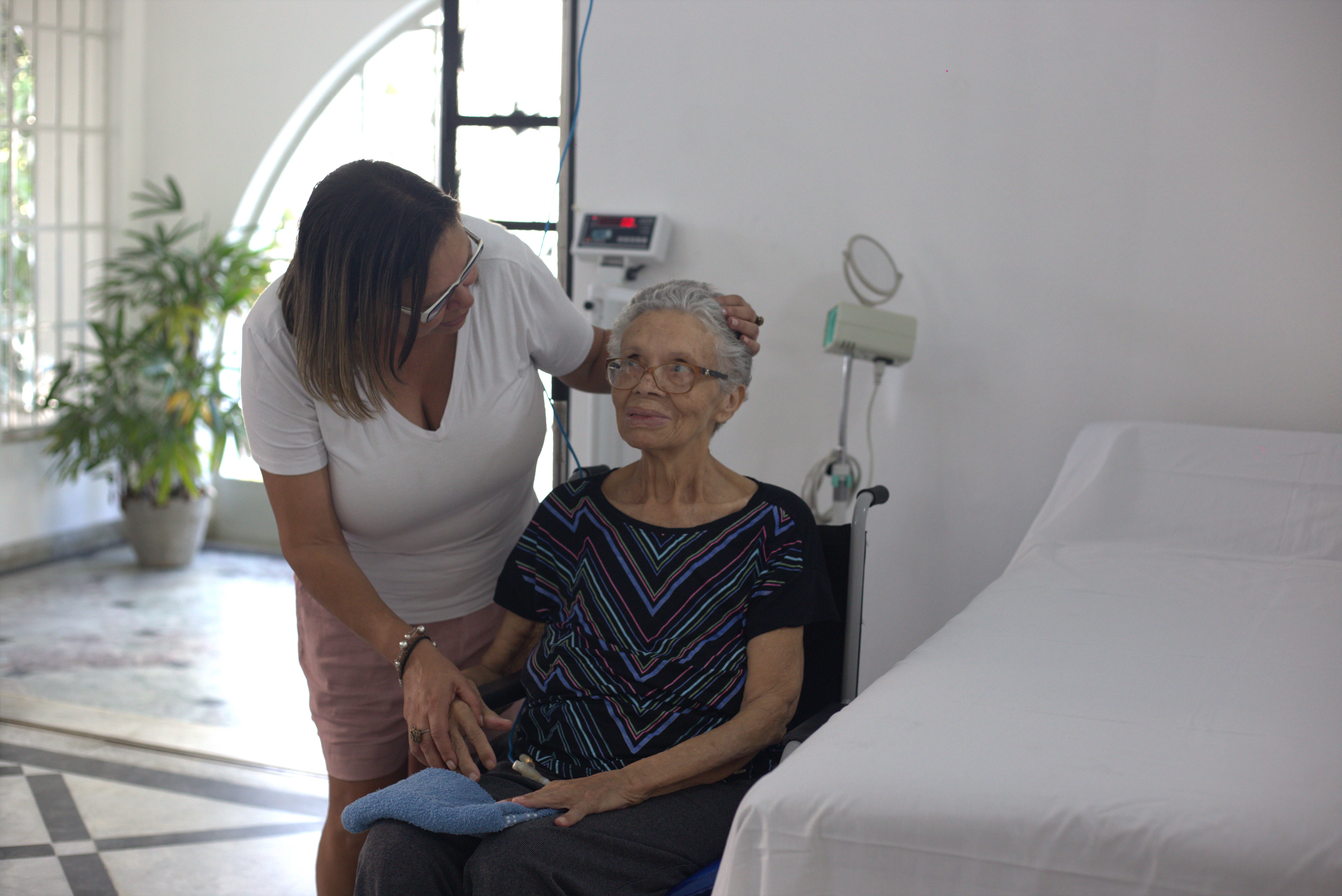 A foto mostra duas mulheres, uma mais jovem e outra idosa; a mulher mais jovem está de pé, levemente reclinada sobre a idosa, sentada em uma cadeira de rodas; uma de suas mãos está pousada sobre a mão da idosa, e a outra pousada em sua cabeça; a idosa sorri e olha para a outra mulher; elas estão ao lado de uma maca, e atrás é possível ver uma balança e um aparelho de medir pressão arterial