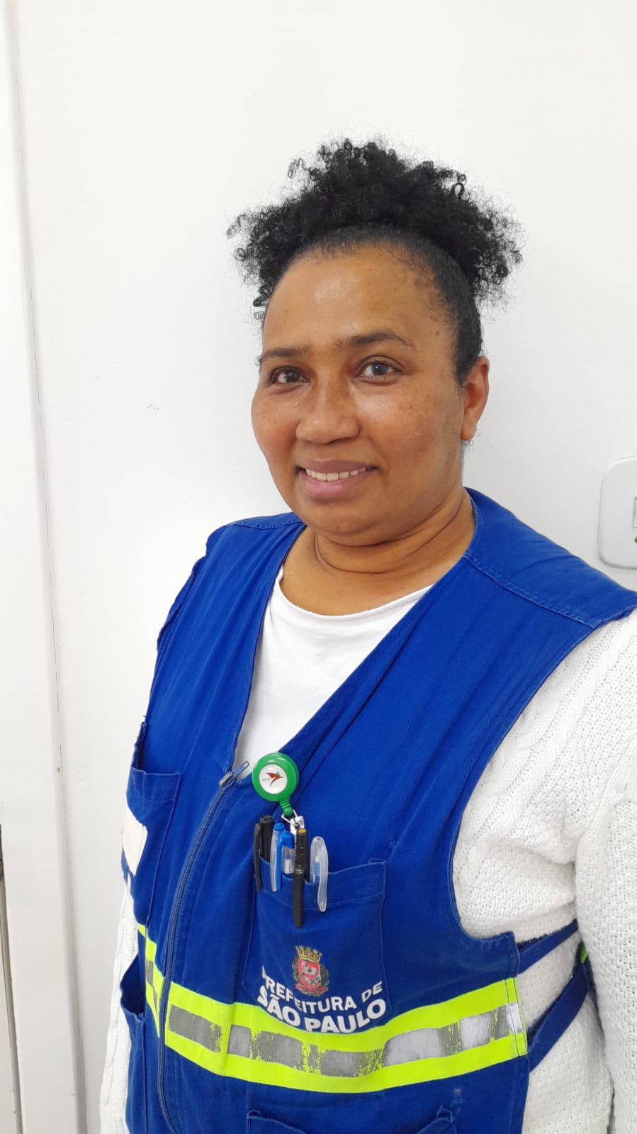 A imagem mostra uma mulher negra de meia-idade; ela está de frente, sorrindo e usa os cabelos e um colete azul com faixa reflexiva amarela e a logomarca da Prefeitura de São Paulo