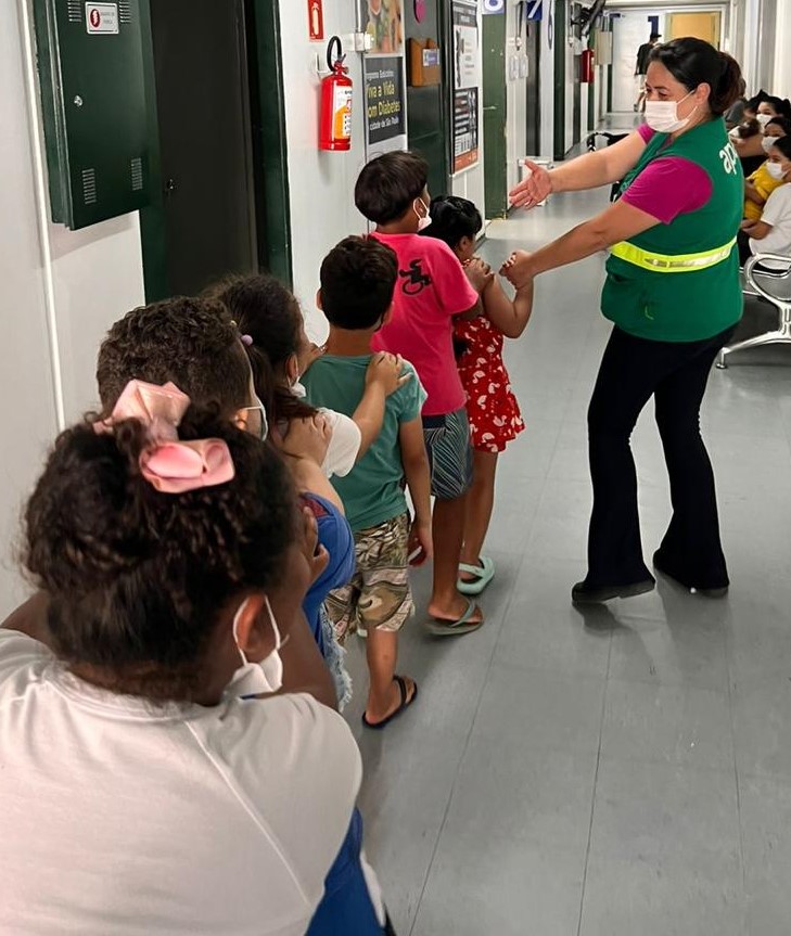 A foto mostra um grupo de crianças sendo conduzido pelo corredor da unidade básica de saúde; elas estão de costas e seguem uma profissional usando um colete verde