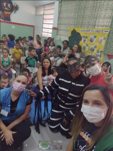 A foto mostra um grupo grande de crianças, sentadas em cadeiras; à frente, estão duas professoras usando máscaras, e dois atores caracterizados como mosquitos, com roupas pretas listradas e máscaras