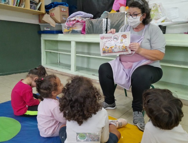 A foto mostra uma professora conversando com crianças pequenas, que estão sentadas à sua frente; a professora segura um pequeno cartaz ilustrado, onde se lê a palavra 