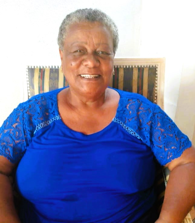 A foto mostra uma mulher negra idosa; ela sorri, tem cabelos curtos e grisalhos, usa uma blusa azul e está sentada