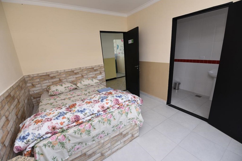 Um dos dormitórios do Serviço Integrado de Acolhida Terapêutica. No canto esquerdo há uma cama de casal com uma colcha florida. É possível ver do lado direito a porta do banheiro aberta