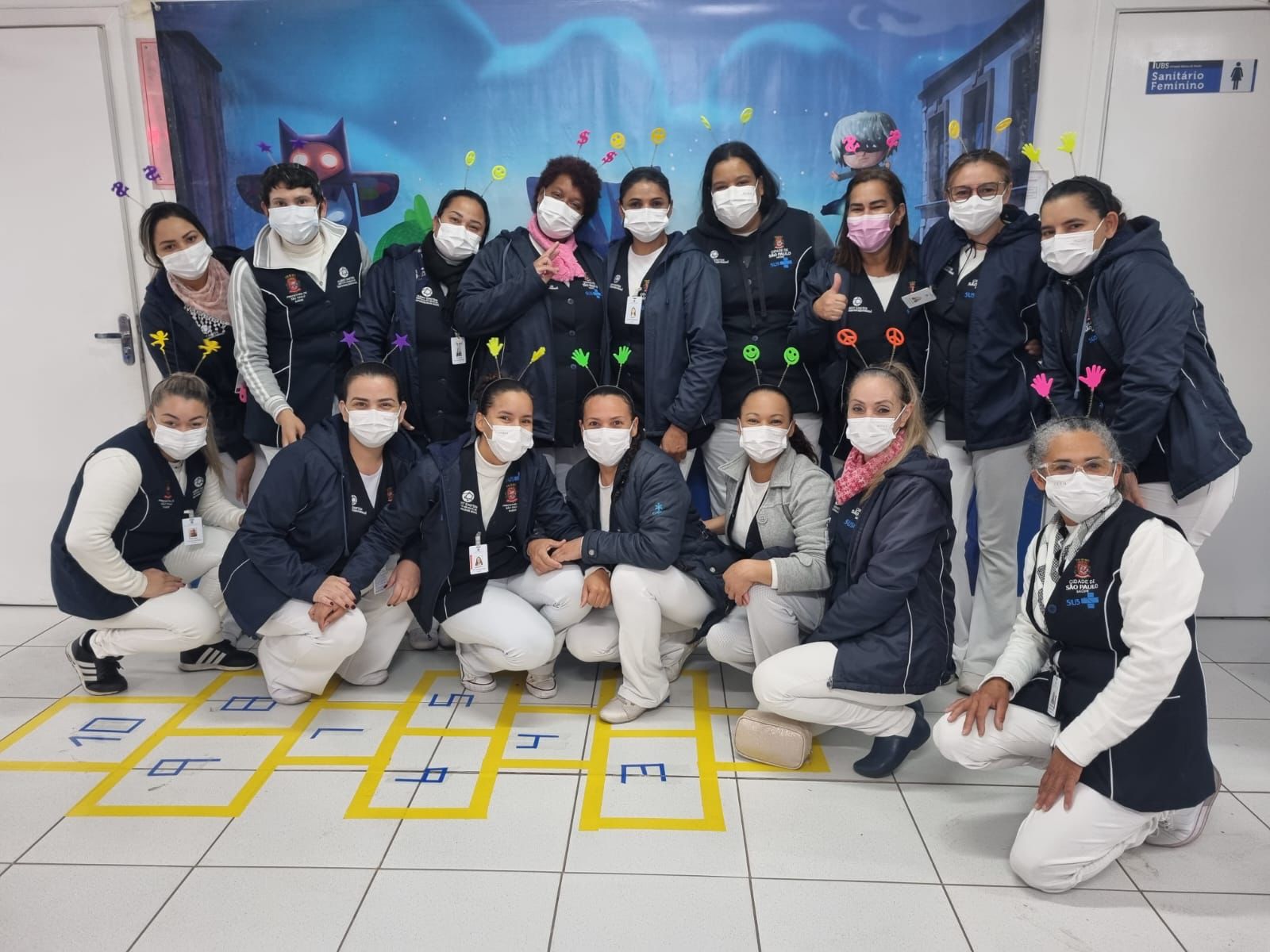 Foto mostra um grupo com 16 mulheres; elas usam uniformes brancos e azuis com a logomarca do Sistema Único de Saúde e o brasão da cidade de São Paulo, usam tiaras com antenas e estão em um ambiente decorado com motivos infantis