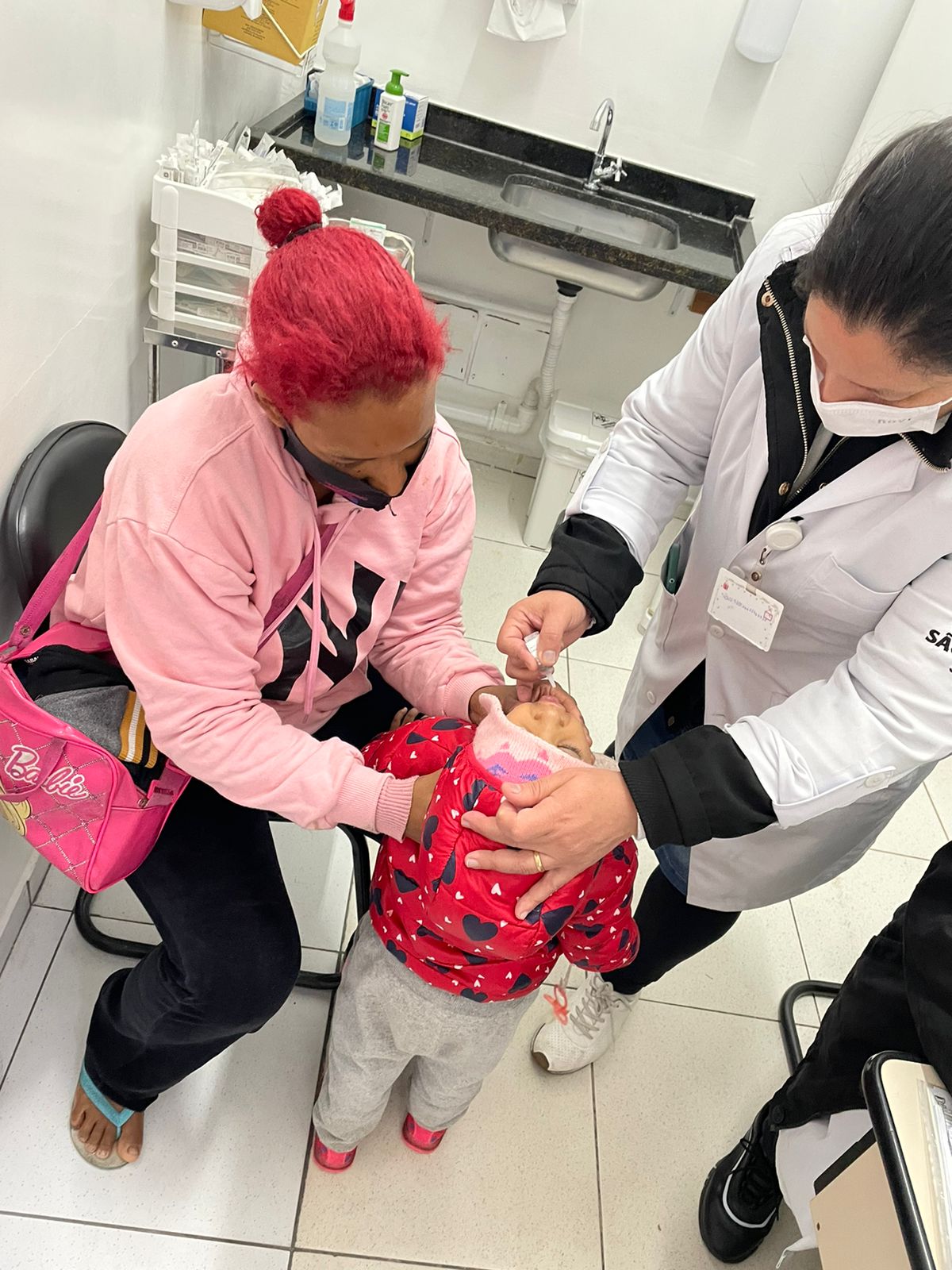 A foto mostra uma criança pequena de costas, com a cabeça levantada, recebendo na boca uma gotinha, aplicada por uma mulher que usa jaleco branco e está posicionada ao seu lado direito; à esquerda, outra mulher apoia a criança com as mãos