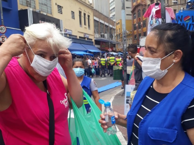 Uma agente de saúde, do lado direito da foto, usando um jaleco azul, ajuda uma mulher que veste camiseta cor de rosa a colocar a máscara. A agente segura na mão um frasco de álcool em gel