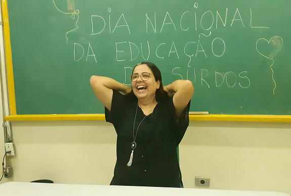 #PraCegoVer: professora Adriana Horta sorri com os braços dobrados na altura da cabeça. Ela está na sala de aula e atrás há um lousa, onde se lê Dia Nacional da Educação de Surdos