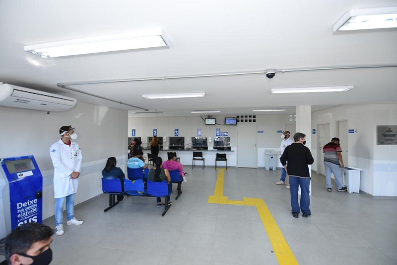 Foto da sala de espera da AMA/UBS Integrada Parque Anhanguera. É possível observar os guichês e pessoas sentadas aguardando atendimento.