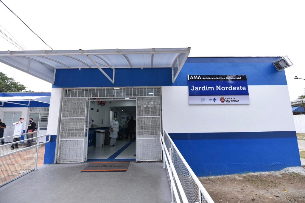 Foto de fachada. Há a entrada de um estabelecimento pintado com cores azul e branco. O portão da entrada é branco e está aberto. Lá dentro, está uma profissional de saúde vestindo branco. Em uma das paredes, há uma placa 