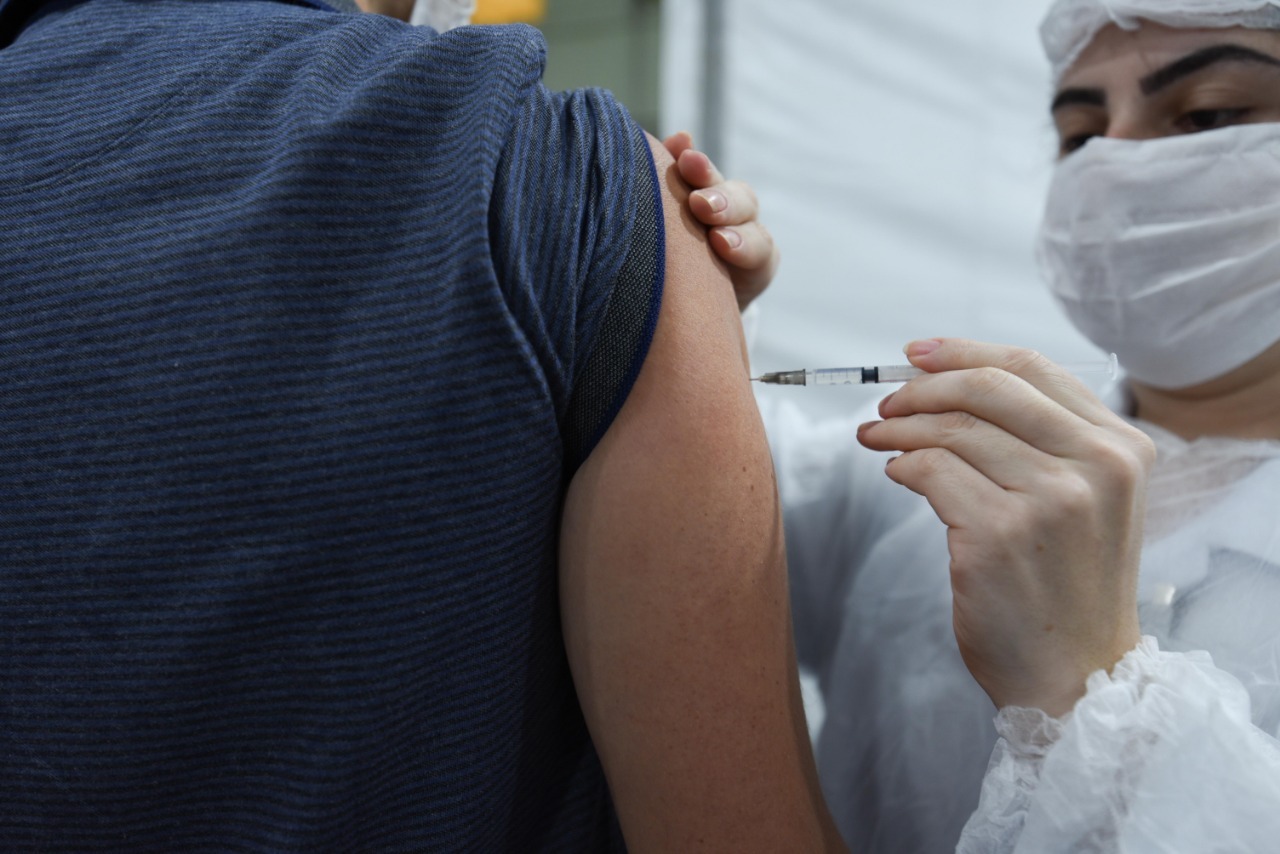 Foto de vacinação. À direita da imagem, uma profissional de saúde de jaleco e máscara de proteção branca está aplicando a dose da vacina em uma pessoa, que está de costas. A pessoa veste uma camisa azul com listras finas pretas.